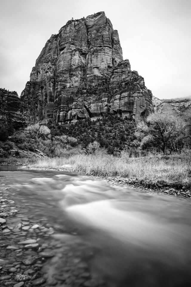 A long-exposure photograph of the Virgin River flowing on the canyon floor of Zion, with Angels Landing in the background.