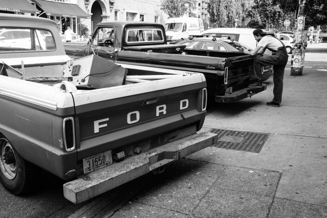 A man in overalls leaning over an old pickup truck parked next to another old pickup truck in Capitol Hill.