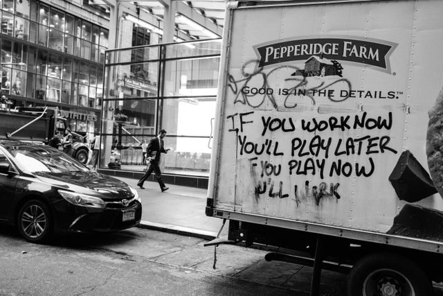 A man walking down the street, next to a Pepperidge Farm truck graffiti'd with "if you work now you'll play later, if you play now you'll work".