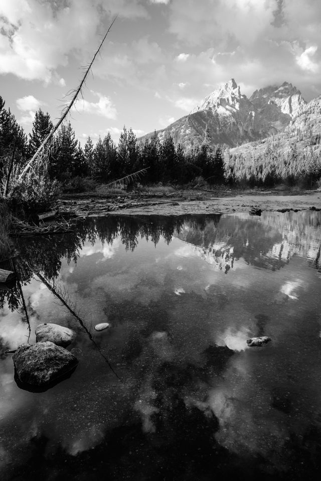 Teewinot Mountain, Grand Teton, and Mount Owen, seen from the outlet of String Lake. Rocks and the reflection of the clouds can be seen in the water.