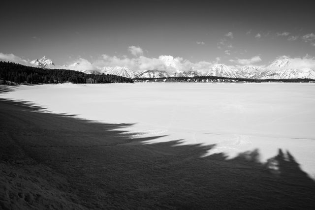 The Tetons, seen from Jackson Lake, which is still drawn down and covered in snow. Its shoreline is lined with shadows of pine trees.