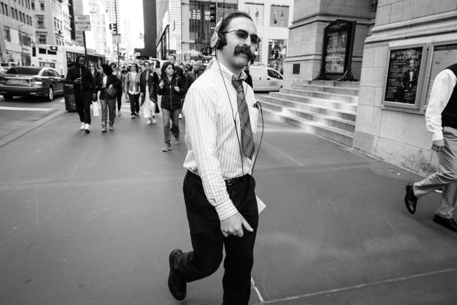 A man with a very bushy mustache wearing sunglasses and headphones smiling at my camera as he walks along Fifth Avenue.