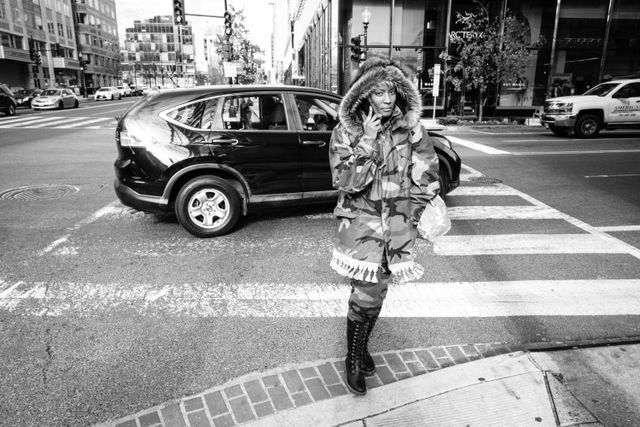 A woman wearing a camo coat crossing the street near CityCenterDC while talking on the phone.