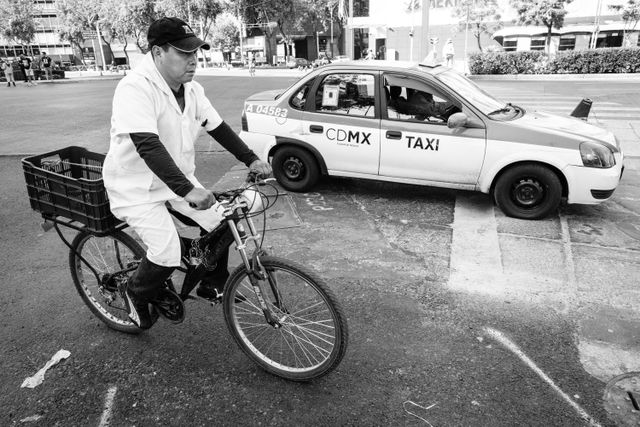 A man in a white coat riding a bike next to a taxi in Mexico City.