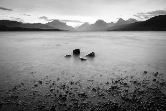 A long exposure photo of a group of four large rocks near the shore of Lake McDonalds during a storm, with the mountains in the background.