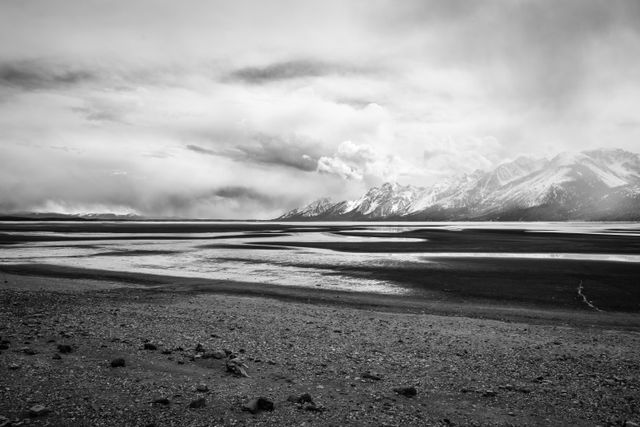 The dry lake of the drawn down Jackson Lake, under stormy skies. In the background, the Teton Range.