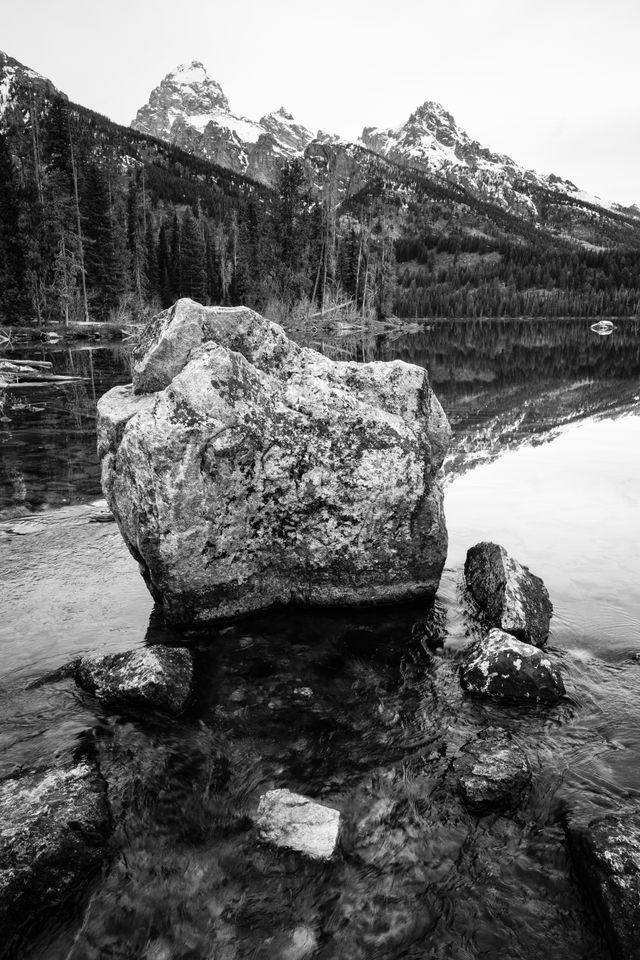 A large boulder in the outlet of Taggart Lake, with water flowing around it. Grand Teton and Teewinot Mountain can be seen in the background.