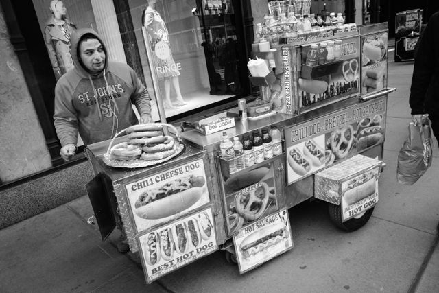 A man in a hoodie working a hot dog stand on Fifth Avenue.