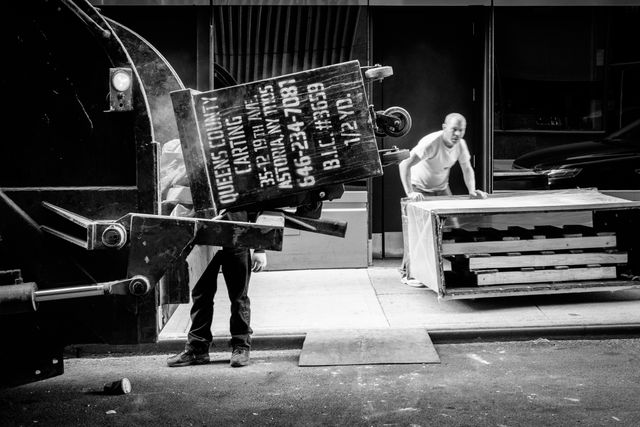 A worker empties a dumpster into a garbage truck while another worker looks on.