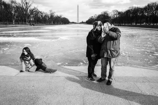 Two people taking a selfie in front of the Lincoln Memorial Reflecting Pool, while another person poses in the background.