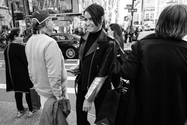 A person in a leather jacket smiling at the camera as they cross 48th Street in New York City.