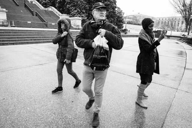 A tourist holding a shopping bag walking on the Capitol grounds as a woman takes a photo.