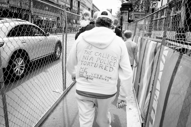 A man walking down Market Street wearing a white hoodie with "the California police tortured me / don't vote for the incumbents please" written in sharpie.