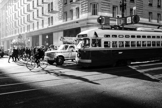 A streetcar lit by morning light on Market Street, with a pickup truck, a person on a bike, and a group of pedestrians next to it.