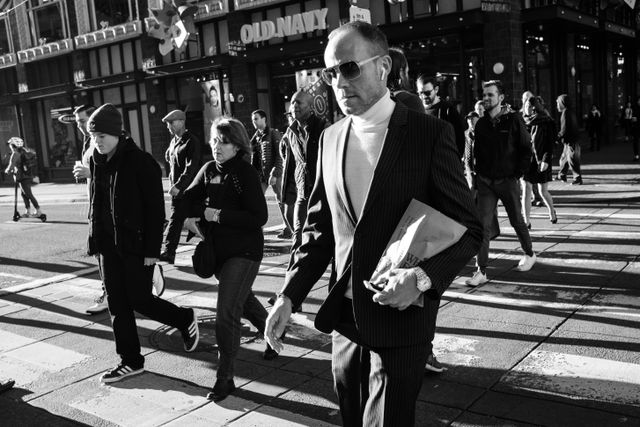 A man wearing a pinstripe suit, a turtleneck, and sunglasses, crossing the street with other pedestrians near Market Street.