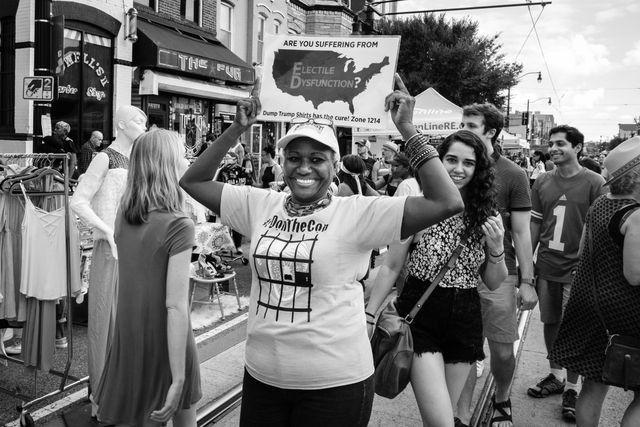 A woman holding up a sign that reads "are you suffering from electile dysfunction?". She's also wearing a "Don the Con" t-shirt.