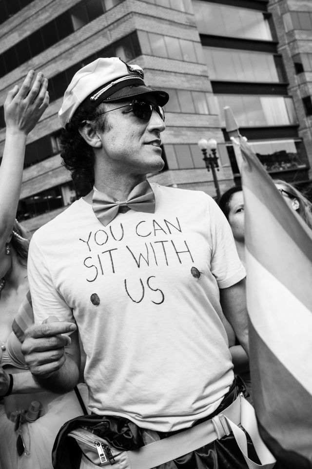 A person at the Capital Pride Parade, wearing a sailor's hat, bowtie, and t-shirt with cutout nipples and "you can sit with us" handwritten on it.