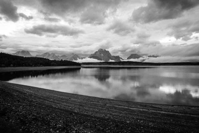 Mount Moran and the Teton Range under stormy skies at dawn. In the foreground, Jackson Lake, with its water level visibly low after a drawdown.