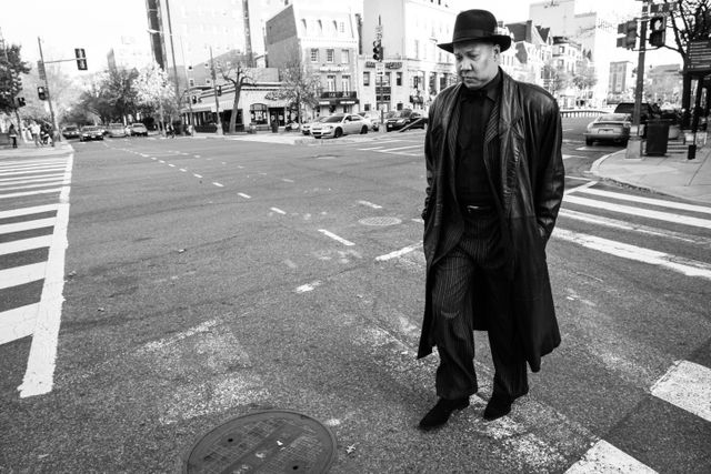 A man wearing a hat, pinstripe suit, and leather overcoat, crossing the street near Dupont Circle.