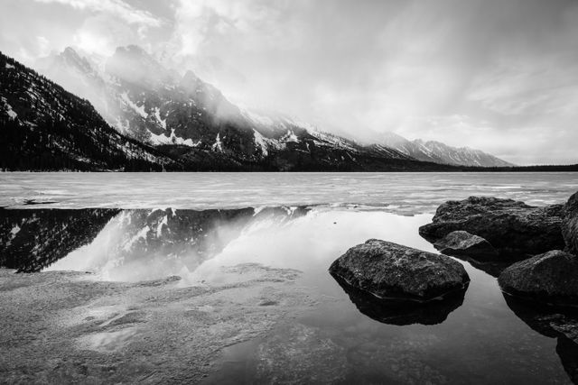 A spring storm clearing over a half-frozen Jenny Lake. In the foreground, boulders on the shore of Jenny Lake; in the background, clouds moving past Storm Point, Symmetry Spire, and Mount Saint John.