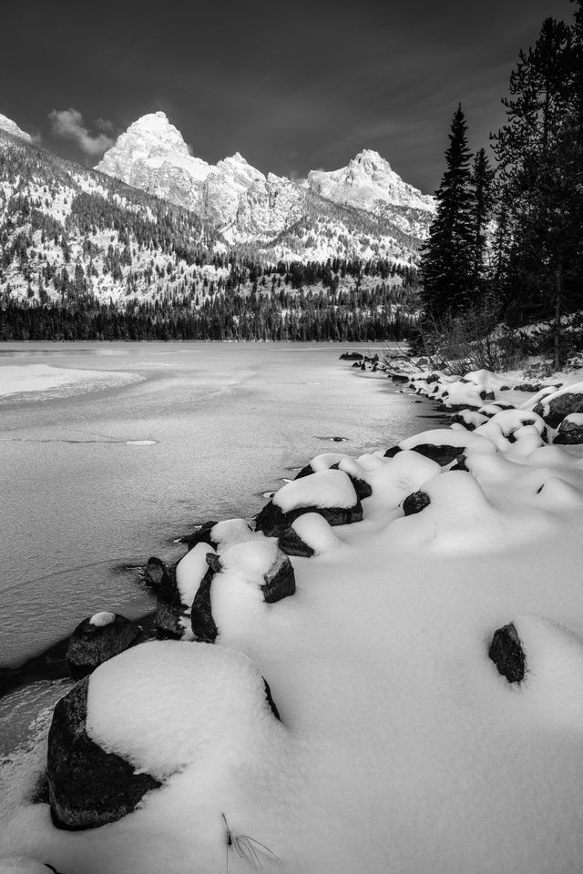 Snow-covered rocks on the shore of a frozen Taggart Lake. In the background, Grand Teton, Mount Owen, and Teewinot Mountain.