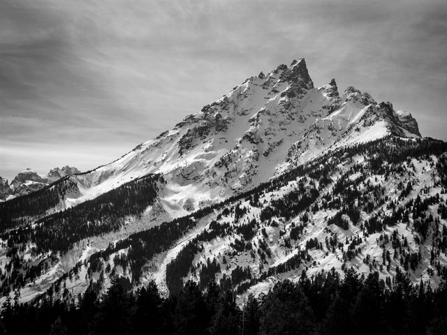 Teewinot Mountain, covered in snow in early spring, under whispy clouds in the afternoon sky.