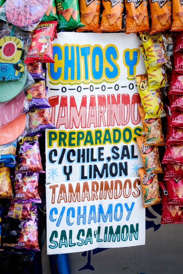 A colorful handpainted sign surrounded by bags of Cheetos, Doritos and other snacks, which reads: "Chitos y tamarindo preparados con chile, sal y limón, tamarindos con chamoy, salsa y limón."