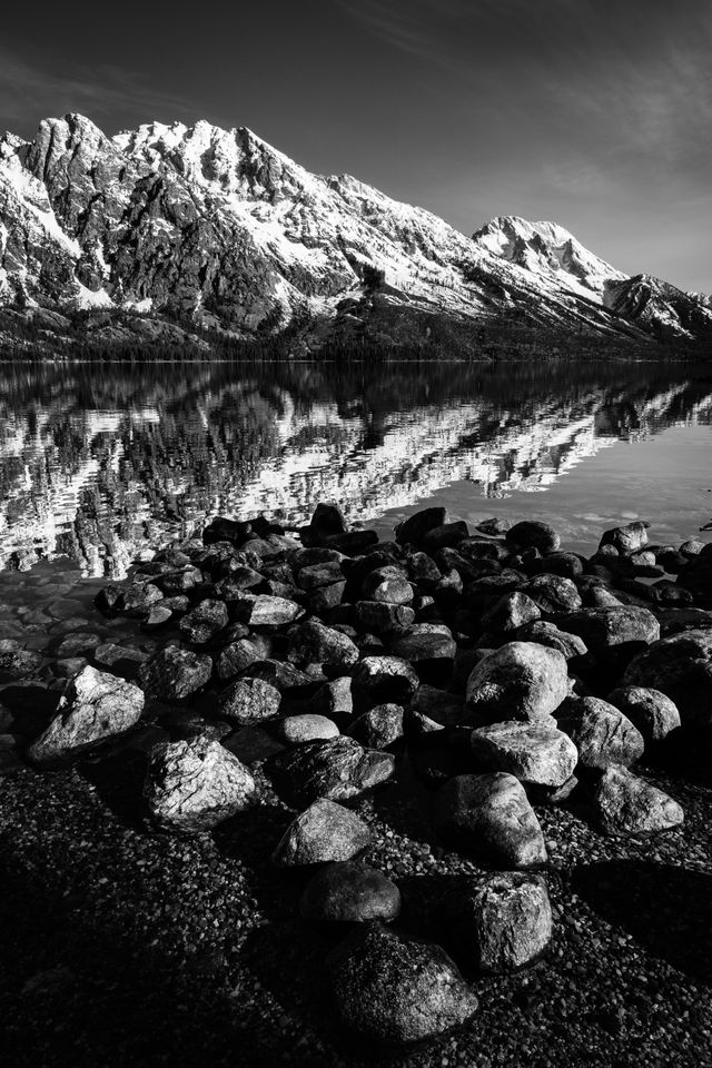 Storm Point, Symmetry Spire, Mount Saint John, and Mount Moran, seen reflected on the surface of Jenny Lake. In the foreground, a group of rocks on the shore of the lake.