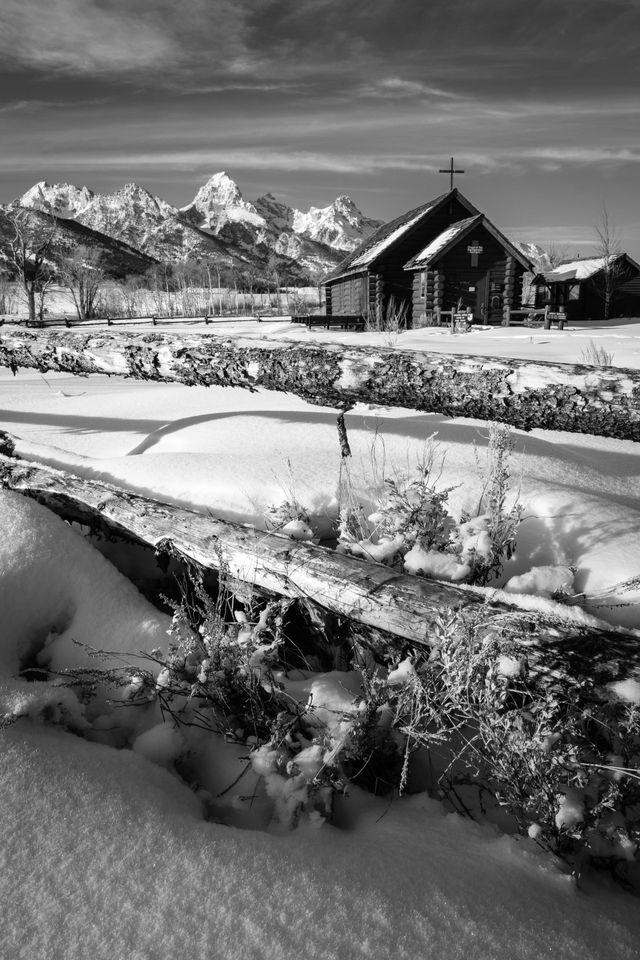 The Chapel of the Transfiguration seen behind a wooden fence in winter, with the Tetons in the background. In the foregound, sagebrush in the snow under the fence.