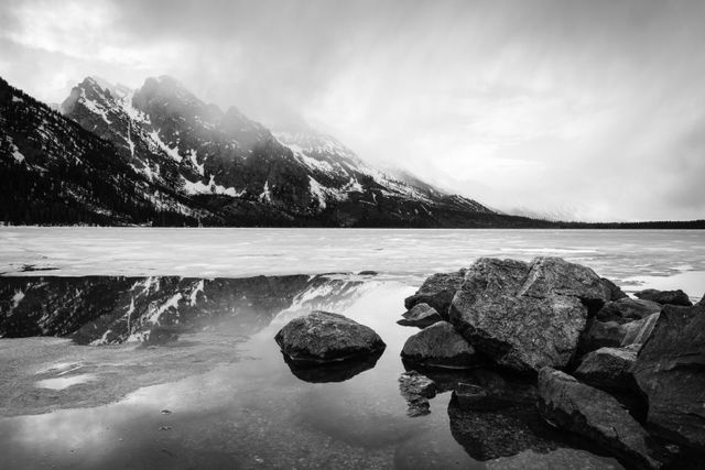 A spring storm moving over a half-frozen Jenny Lake. In the foreground, boulders on the shore of Jenny Lake; in the background, clouds moving over Storm Point, Symmetry Spire, and Mount Saint John.