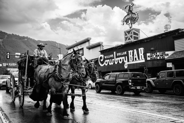 A man riding a horse-drawn stagecoach on a wet street. The stagecoach is pulled by two horses. In the background, there is a building with a sign that reads "Million Dollar Cowboy Bar" and several parked vehicles. The bar has a rotating sign of a man riding a bucking horse with the text "Cowboy Bar". A mountainous landscape can be seen in the distance.