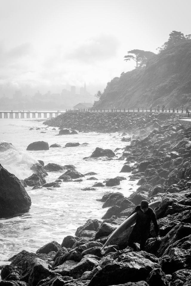 A surfer walking on the rocks in front of Fort Point, with the San Francisco skyline in the background.