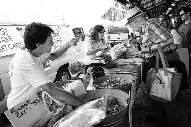 A man wearing a hat shopping for vegetables at Eastern Market while a salesperson fills a bag with leafy greens and another weighs a tomato.