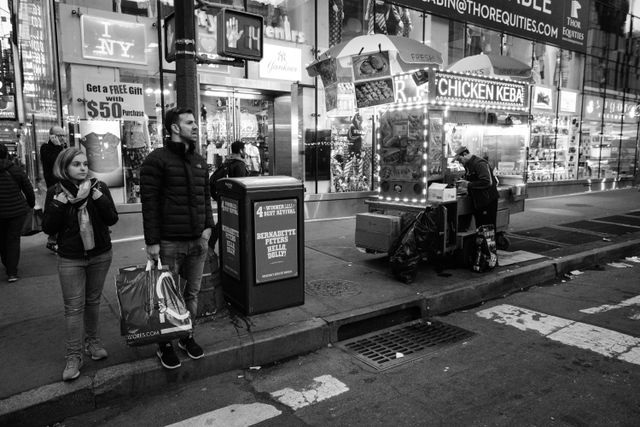 People waiting to cross Eighth Avenue next to a fast food cart.