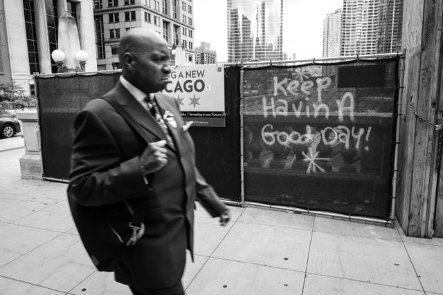 A frowning man walking in front of a graffito that reads "keep havin a good day!" in Chicago.