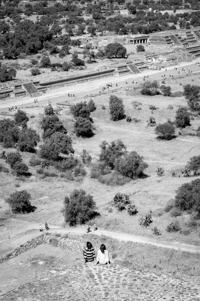 Two tourists sitting at the edge of the Pyramid of the Sun in Teotihuacán, with the Avenue of the Dead in the distance.