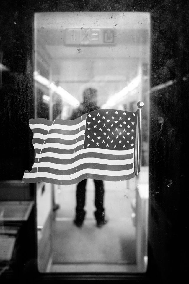 An American flag on the window of the door at the end of a Metro car, with a man standing on the next car in the background.