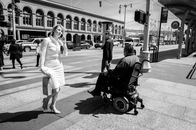 A woman dressed in white walking past a man in a wheelchair in front of the Ferry Building in San Francisco.