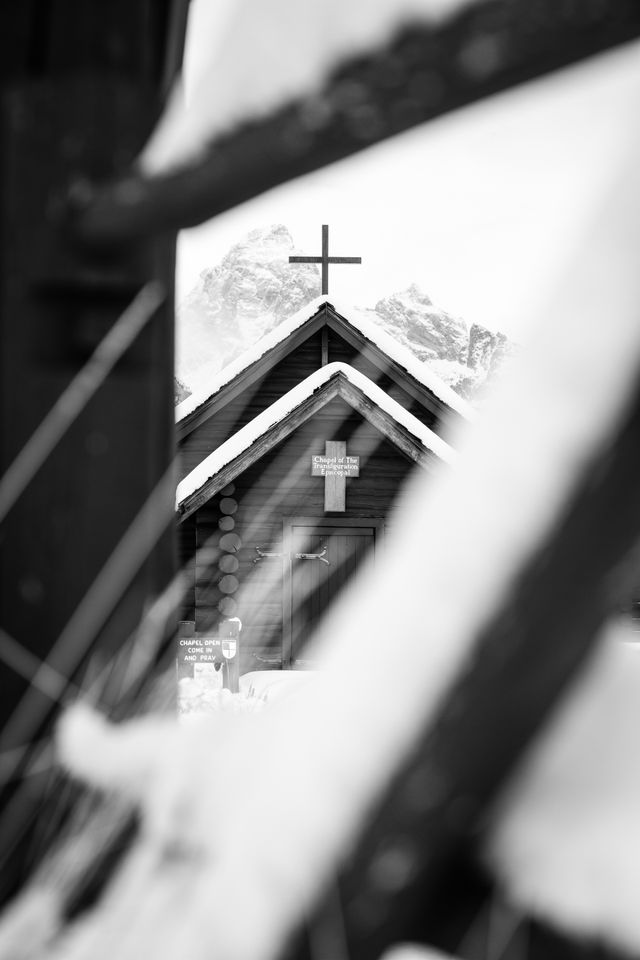 The Chapel of the Transfiguration, seen through a snow-covered fence. The roof of the chapel is covered in snow. In the background, Grand Teton and Mount Owen, under gray skies. A sign above the front door reads "Chapel of the Transfiguration Episcopal", and another to the left of the front door reads "chapel open, come in and pray".