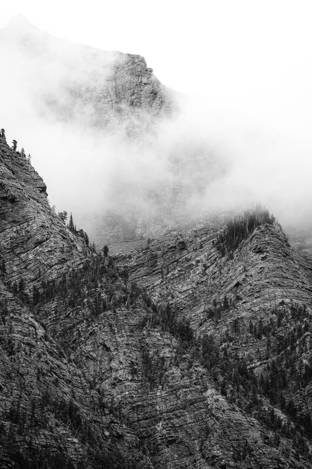 Clouds hanging on the side of Bearhat Mountain, as seen from Avalanche Lake. The mountain is striated and sparsely covered in trees.