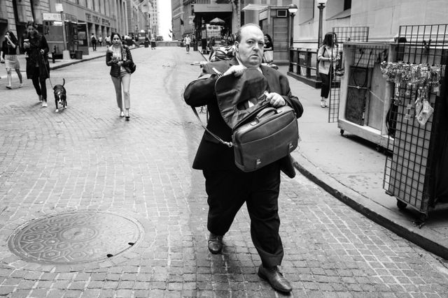 A man in a suit walking on Wall Street while trying to open his suitcase.