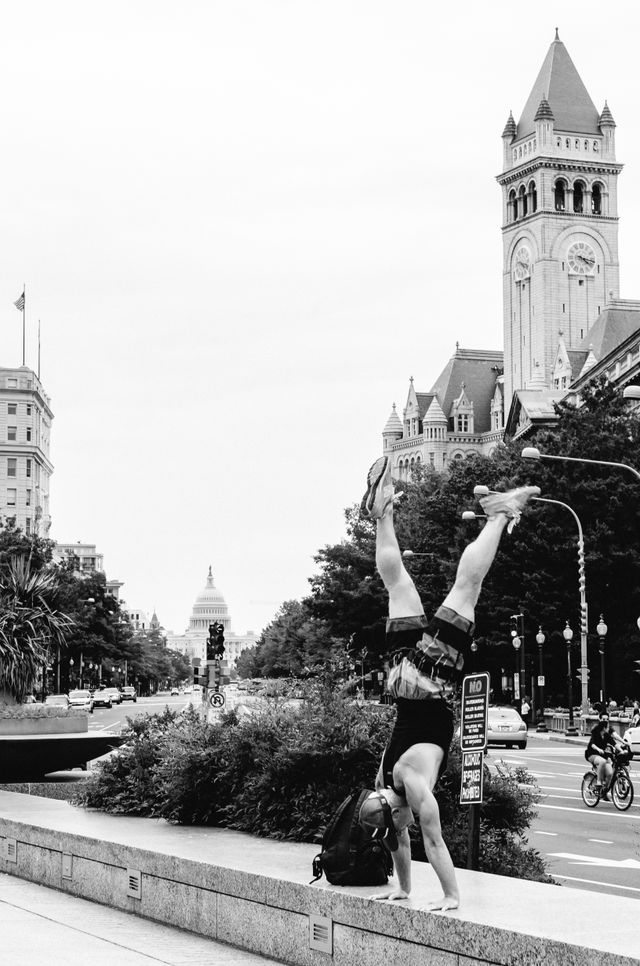 A man doing a head stand at Freedom Plaza in Washington, DC, with the Old Post Office building and the United States Capitol in the background.