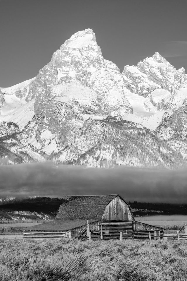 A classic view of the John Moulton barn in Mormon Row, in front of a snow-covered Grand Teton. A layer of clouds and fog hangs just above the barn.