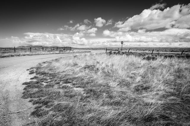 A curving dirt path leading to a paved road with a stop sign. A wooden fence is along the left side of the dirt road; the paved road has barbed wire fences on its sides. Wind-blown grasses are in the foreground; the sky is partly cloudy, and a snowstorm can be seen behind a hill in the distance.