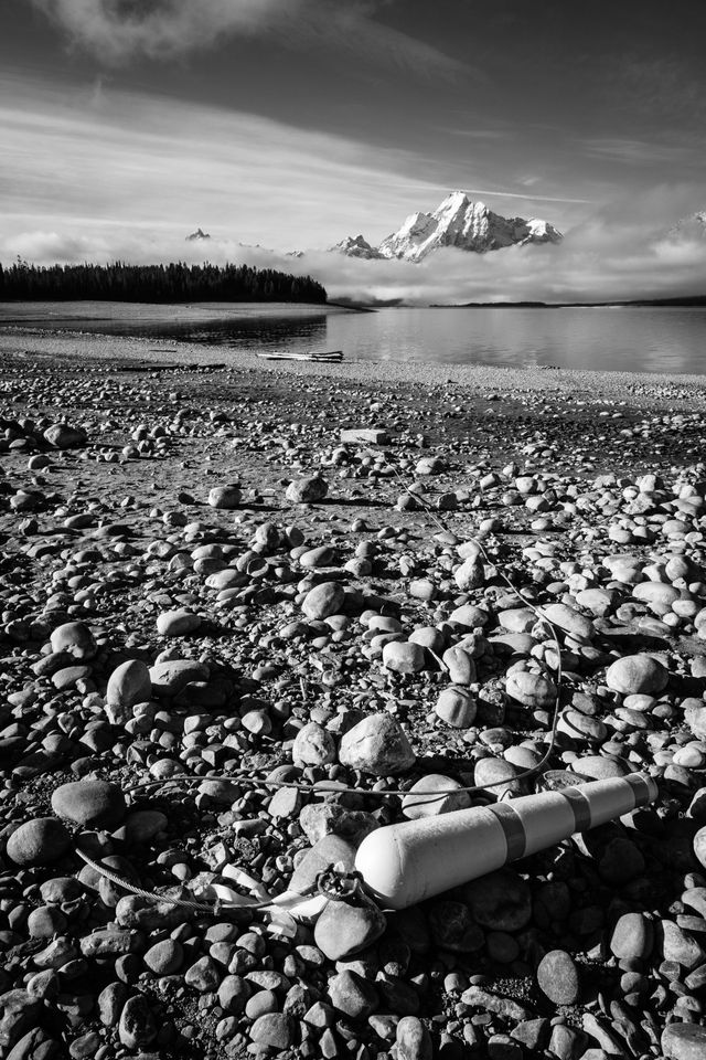 A buoy laying on the dry lakebed of Jackson Lake, along with its steel wire and concrete anchor. In the background, Mount Moran and Grand Teton, partially covered in clouds.