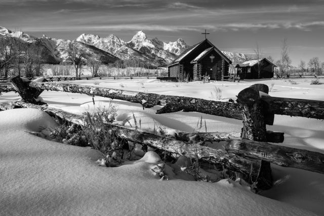 The Chapel of the Transfiguration seen behind a wooden fence in winter, with the Tetons in the background. In the foregound, sagebrush in the snow under the fence.