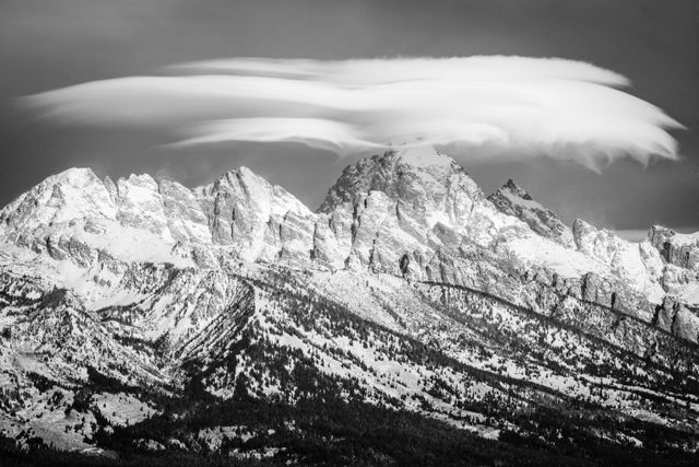 The Teton range, including Nez Perce Peak, Grand Teton, and Mount Owen, covered in snow with a whispy lenticular cloud hanging over the summit of The Grand. Snow can be seen blowing off the top of some of the peaks.