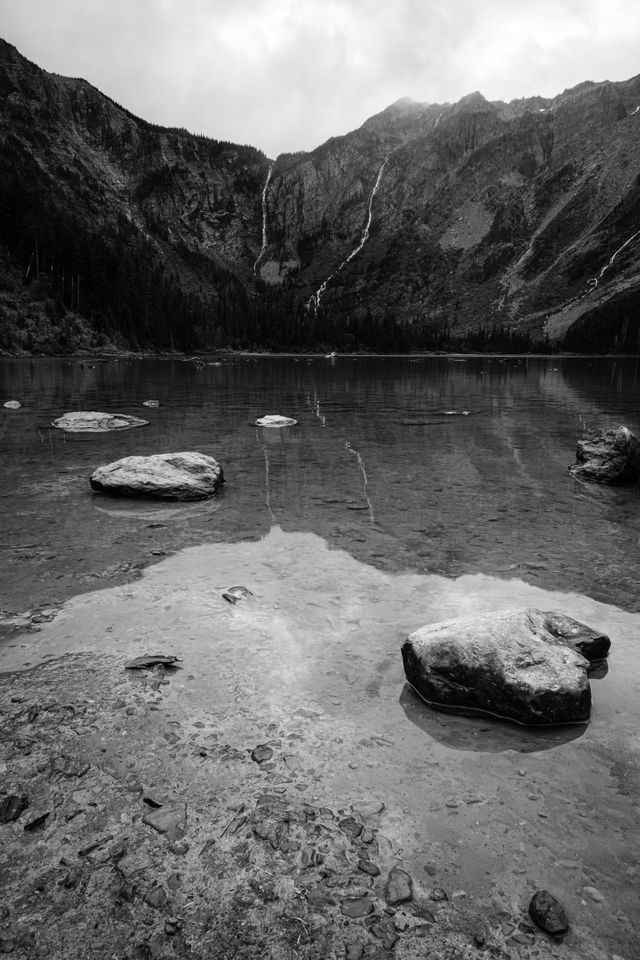 Avalanche Lake. In the foreground, a group of seven large half-submerged boulders near the shore of the lake, and in the background, three waterfalls running down the mountains at the end of the lake.