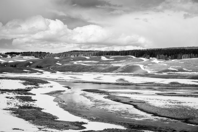 The Yellowstone River in Yellowstone National Park. Its banks are covered on snow, and in the background, rolling hills and a line of trees can be seen.