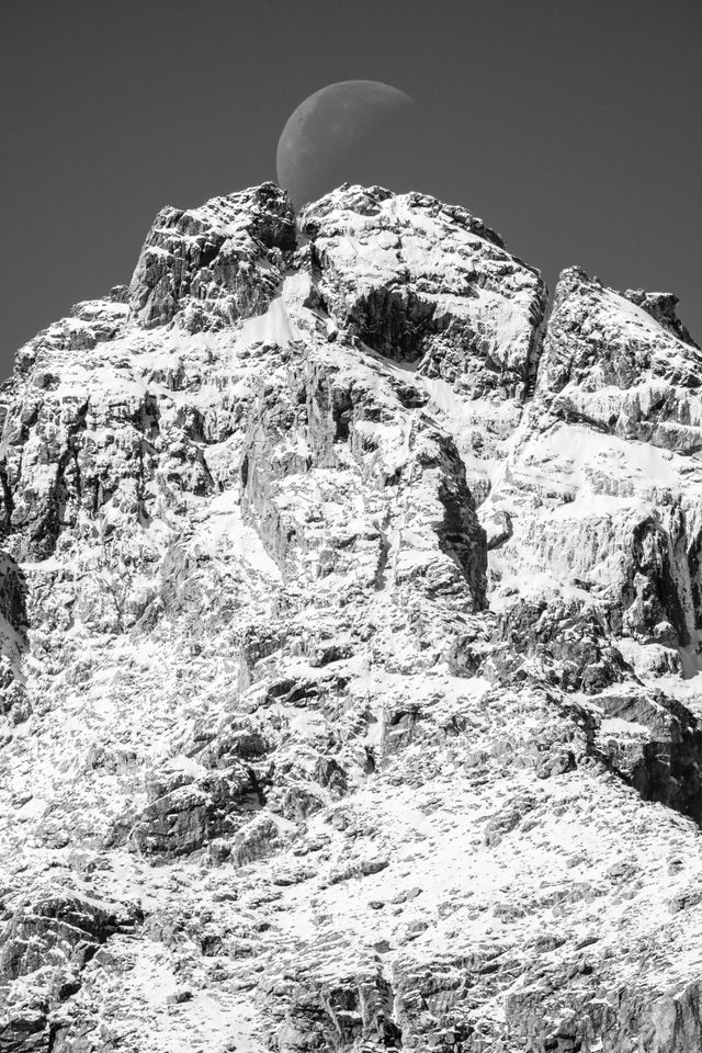 A waning crescent moon, setting directly behind the summit of a snow-covered Middle Teton.